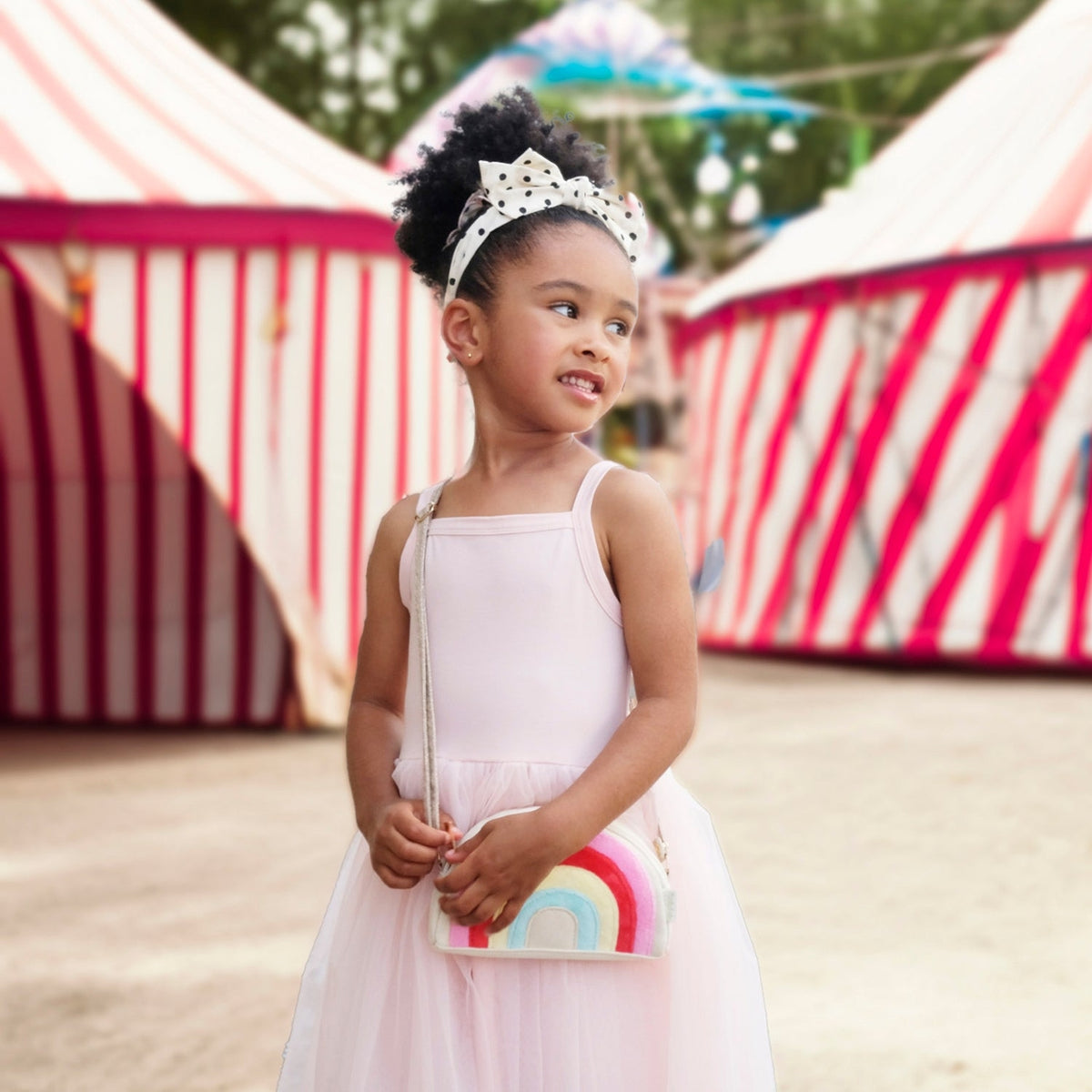 Young girl in a white dress with a rainbow clutch standing in front of striped tents.