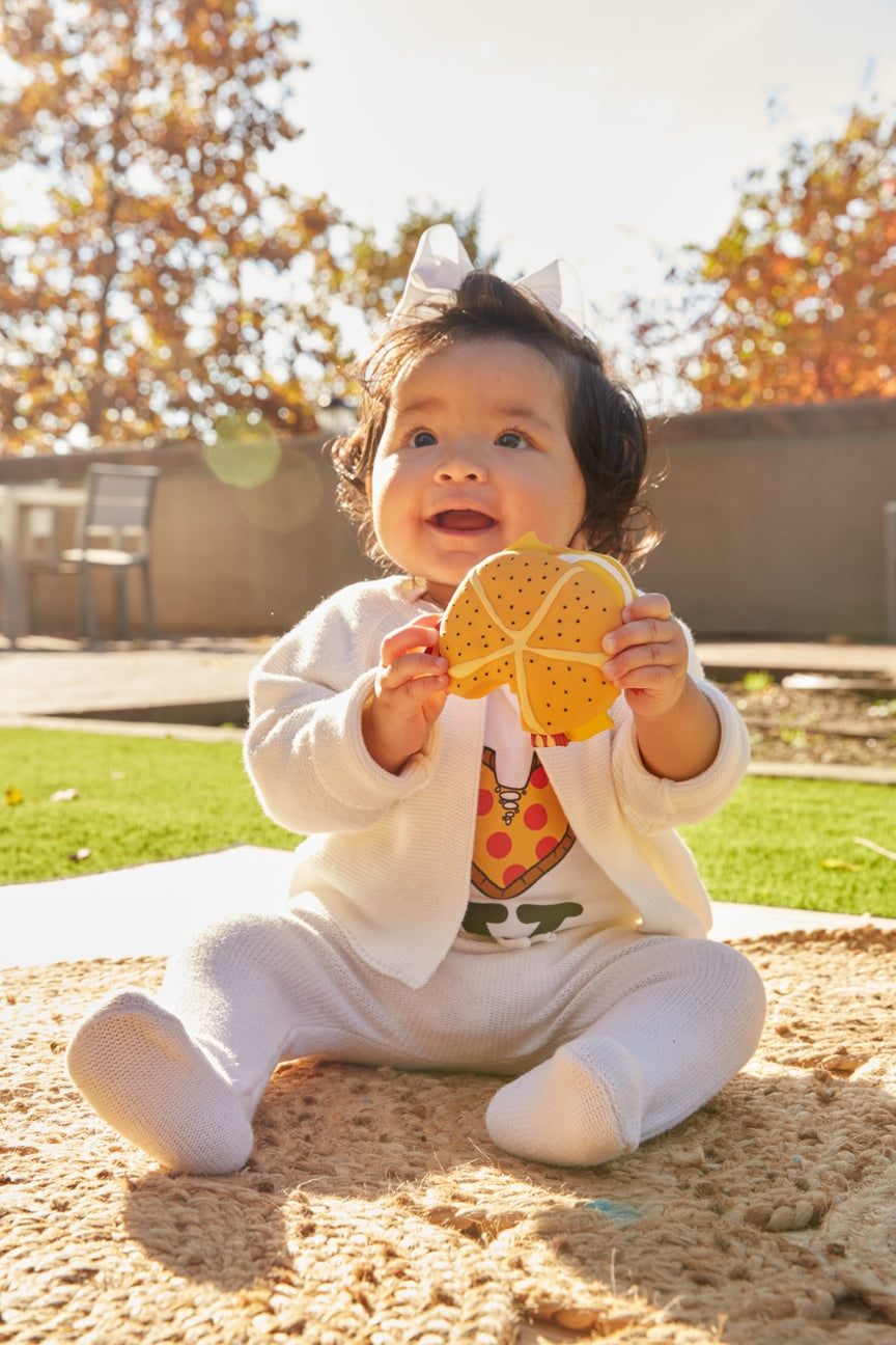 A baby sitting outdoors on a sandy surface wearing a white outfit and holding a yellow and orange patterned toy, with a blurred garden background.