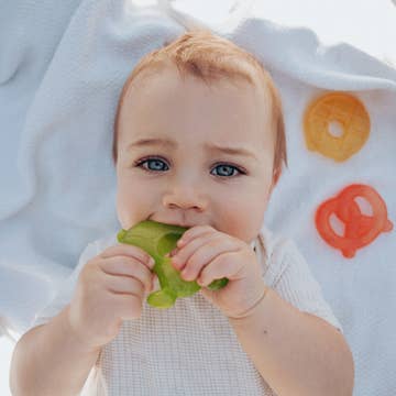 Baby chewing on a green toy with a white blanket and orange shapes in the background