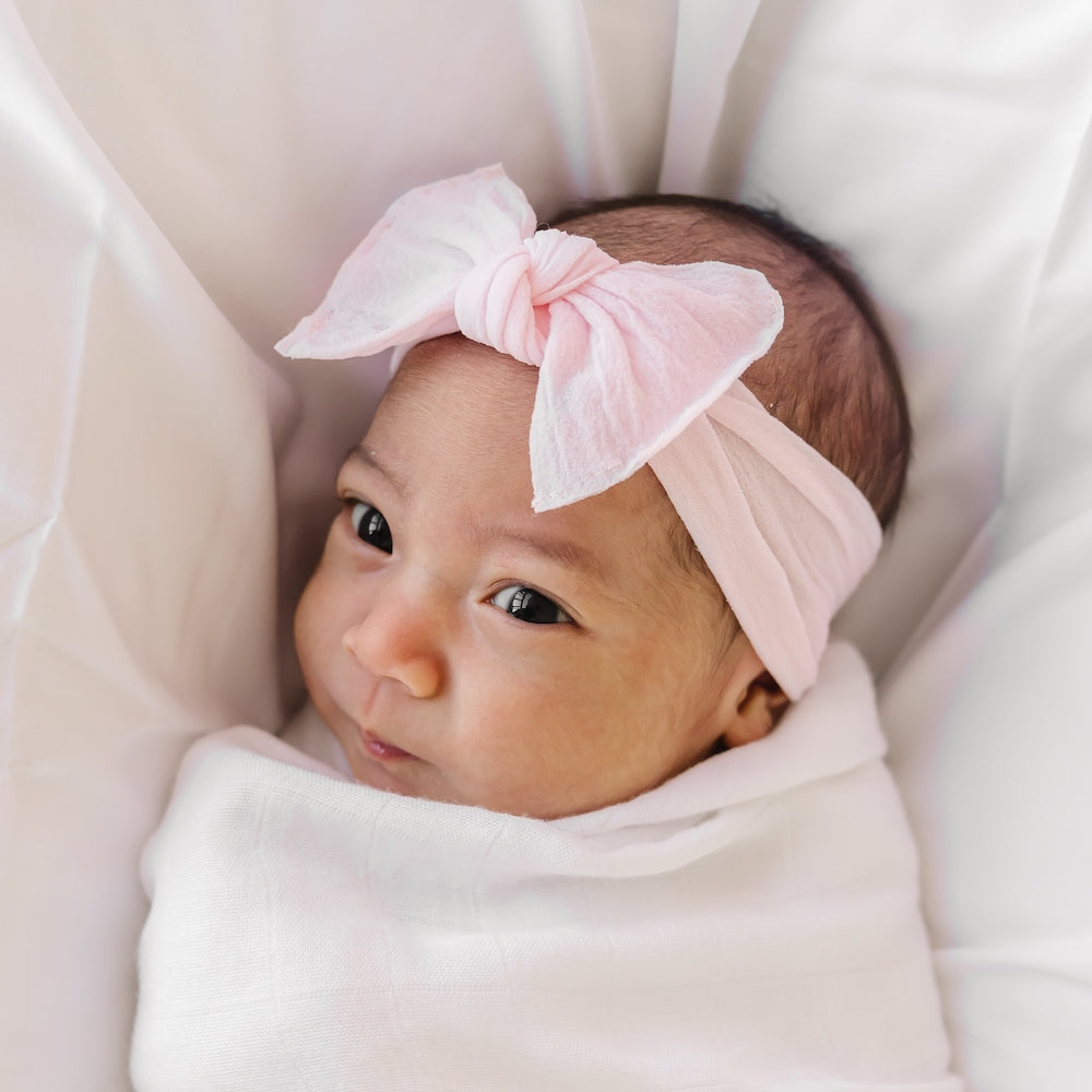 Newborn baby wearing a pink headband with a bow, wrapped in white fabric.