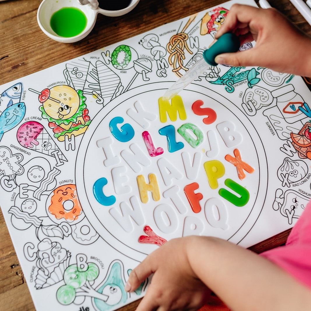 Coloring book with alphabet letters and children's drawings on a wooden table.