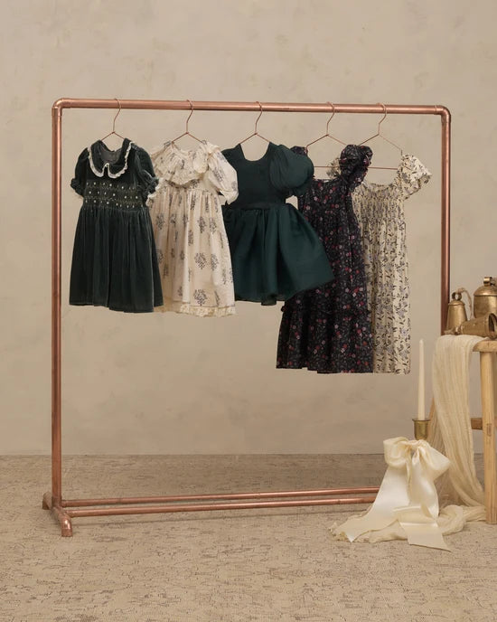A copper-colored clothing rack displaying five vintage-style children's dresses in various colors and floral patterns, set against a neutral textured beige wall and floor.