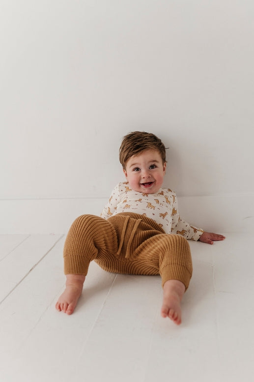 A baby sitting on a white floor against a white wall, wearing a cream long-sleeve top with a small tan pattern and ribbed mustard-brown pants.