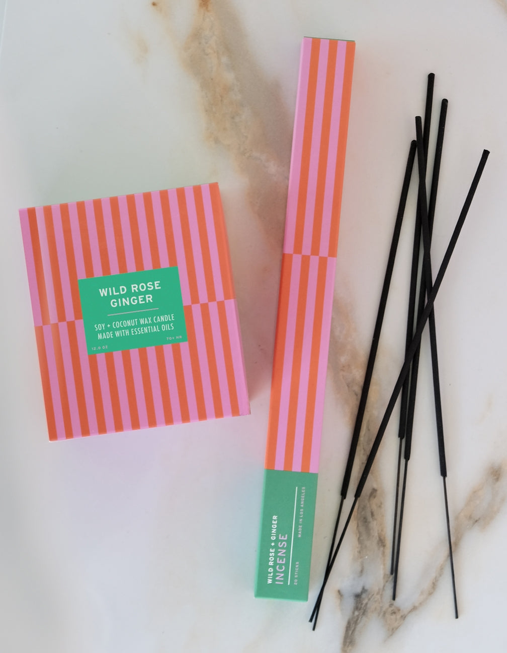 A flat-lay of Wild Rose Ginger incense, showing the pink and orange striped box, the incense stick package with a green label, and several black incense sticks on a white marble background.