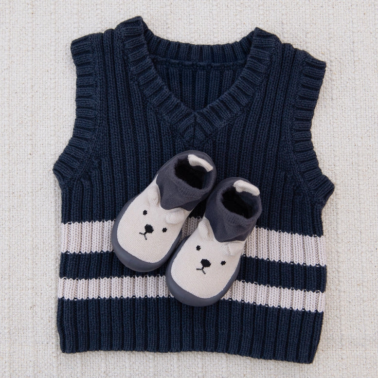 A baby knit sweater vest in navy blue with two horizontal white stripes and a white bootie with a navy cuff and embroidered bear face, laid flat on a textured beige fabric background.