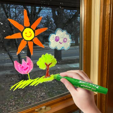 Hand holding a green window gel crayon with colorful window decorations on a glass door.