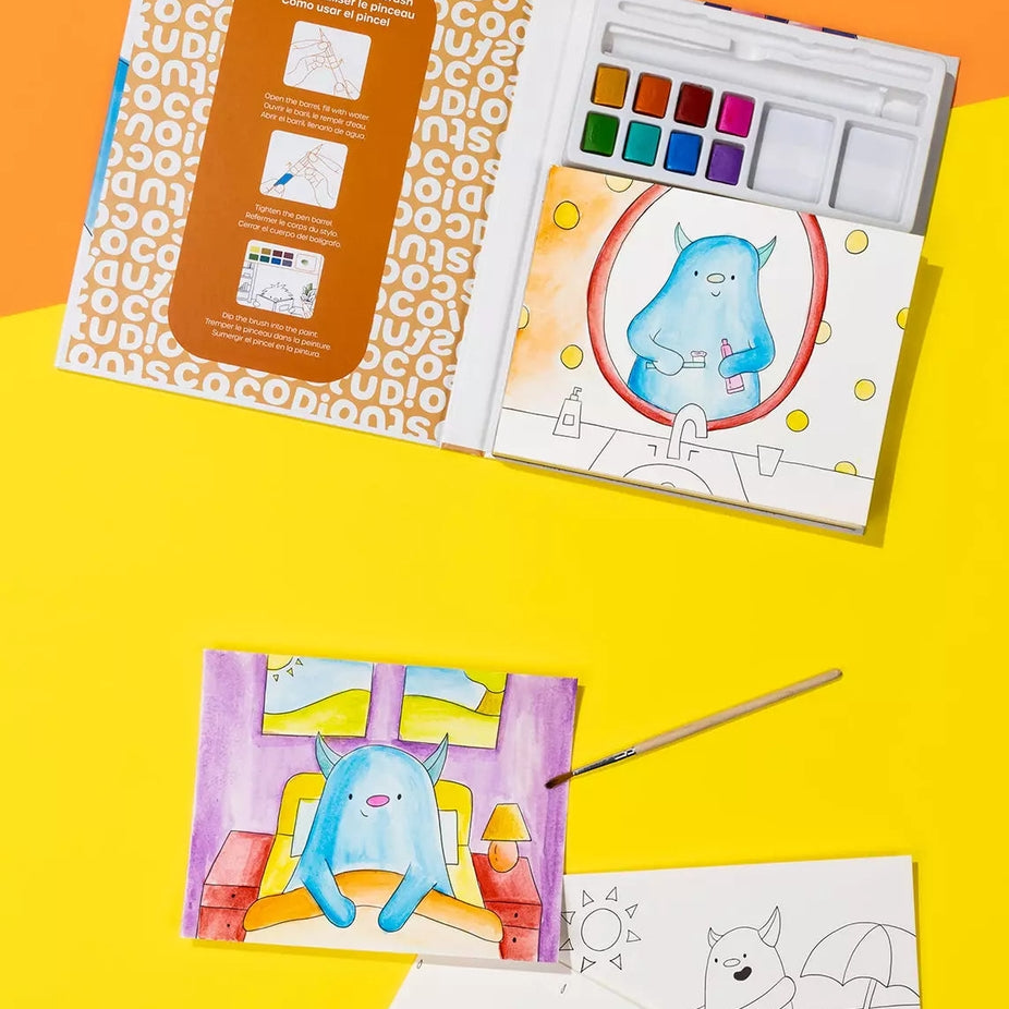 A children's watercolor painting kit shown open on a bright yellow and orange background, with coloring pages, a paintbrush, and a watercolor palette visible. The kit includes printed coloring pages and a painted illustration of a blue monster.