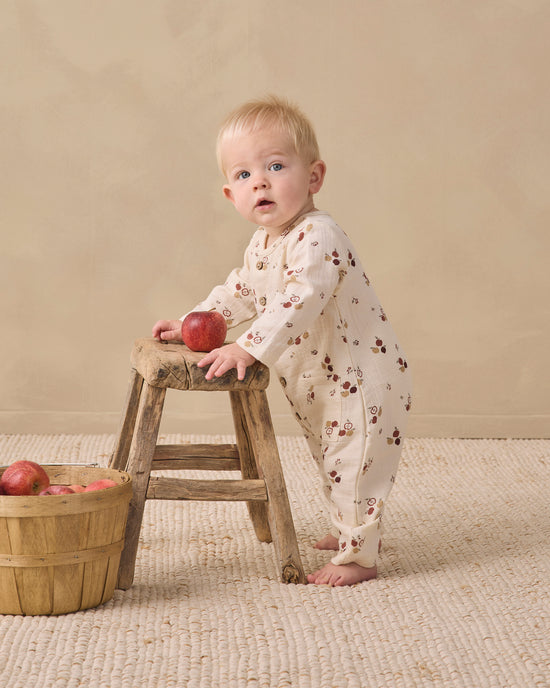 A baby standing on a woven rug and leaning on a rustic wooden stool, wearing a cream footed sleeper with a brown cherry print. A small wooden basket with red apples sits on the floor to the left.