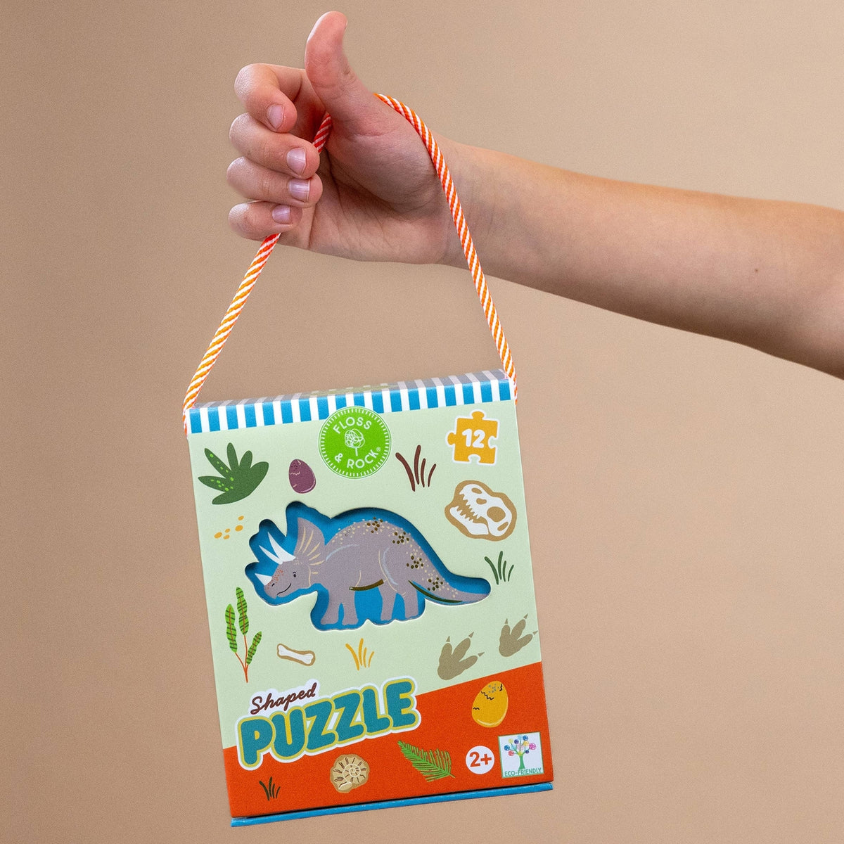 Hand holding a dinosaur-themed shaped puzzle packaging against a beige background
