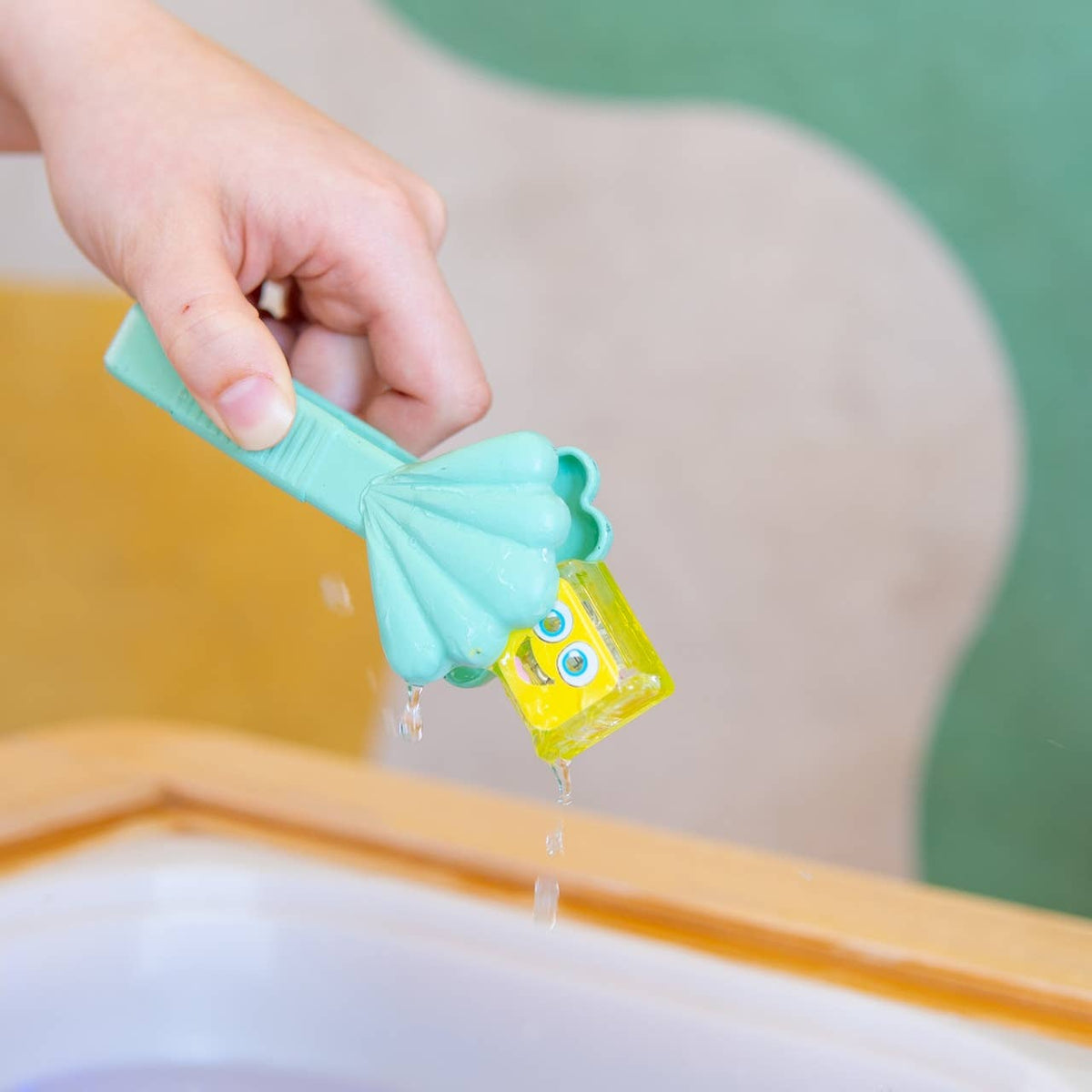 Hand holding a green and yellow tool with water dripping from it, against a blurred background.