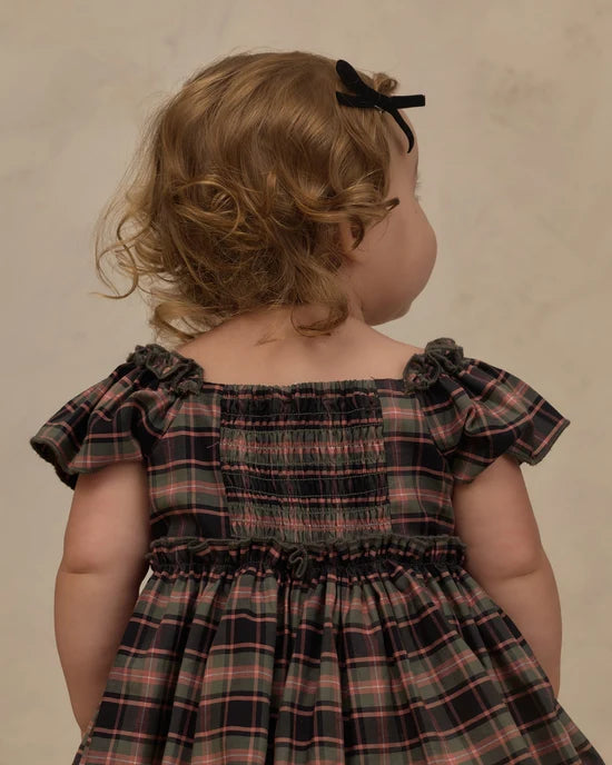 A toddler viewed from behind wearing a smocked plaid dress in dark green, red, and black, with a black bow in her hair, set against a neutral textured beige background.