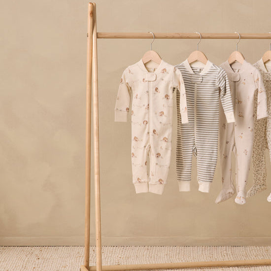 Four baby footed sleepers in neutral tones hanging on a wooden clothing rack against a textured beige wall and floor.