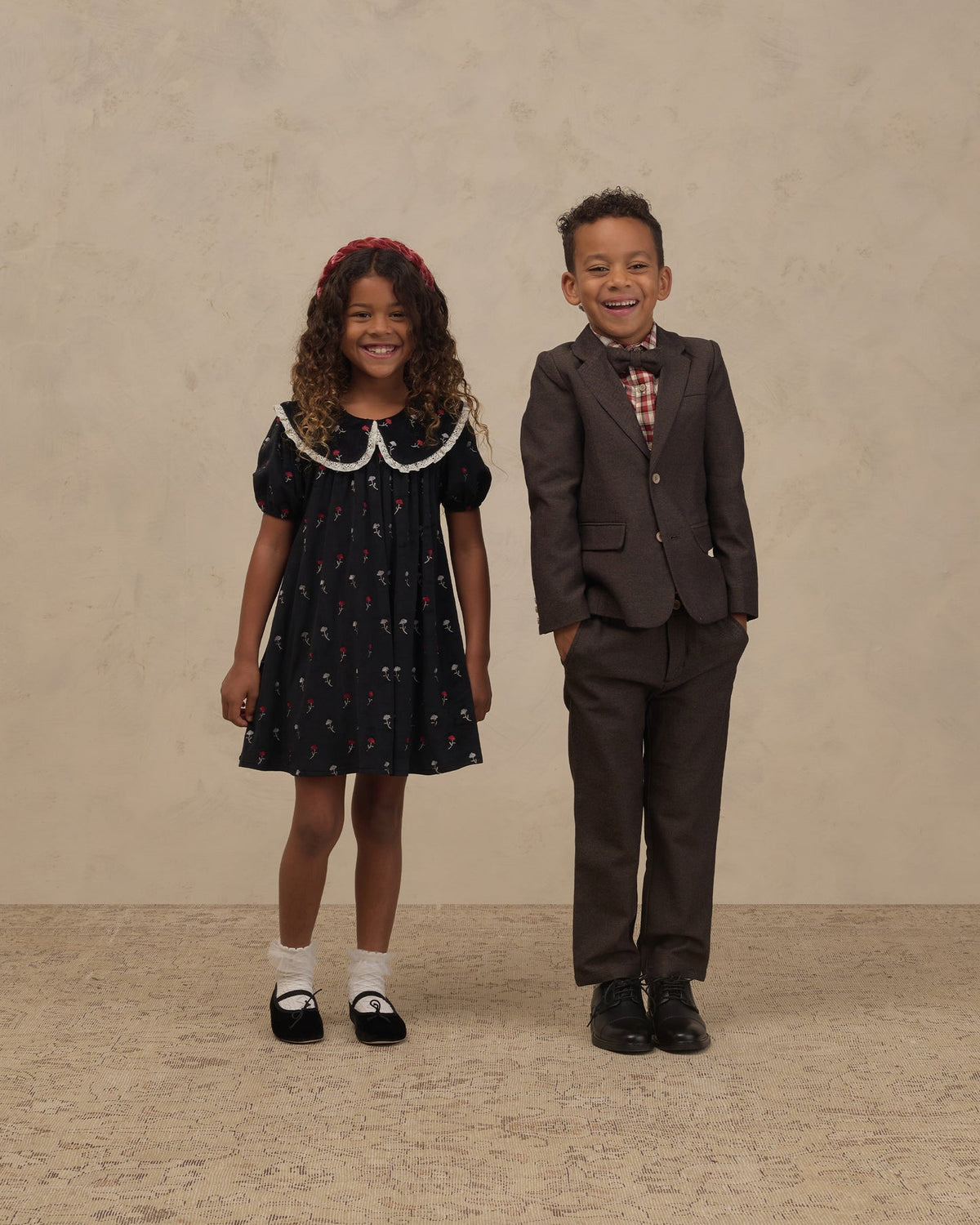 A young girl in a dark navy floral dress with a white Peter Pan collar and a red headband stands next to a young boy in a dark brown suit, bow tie, and dress shirt, posing against a textured beige studio backdrop.