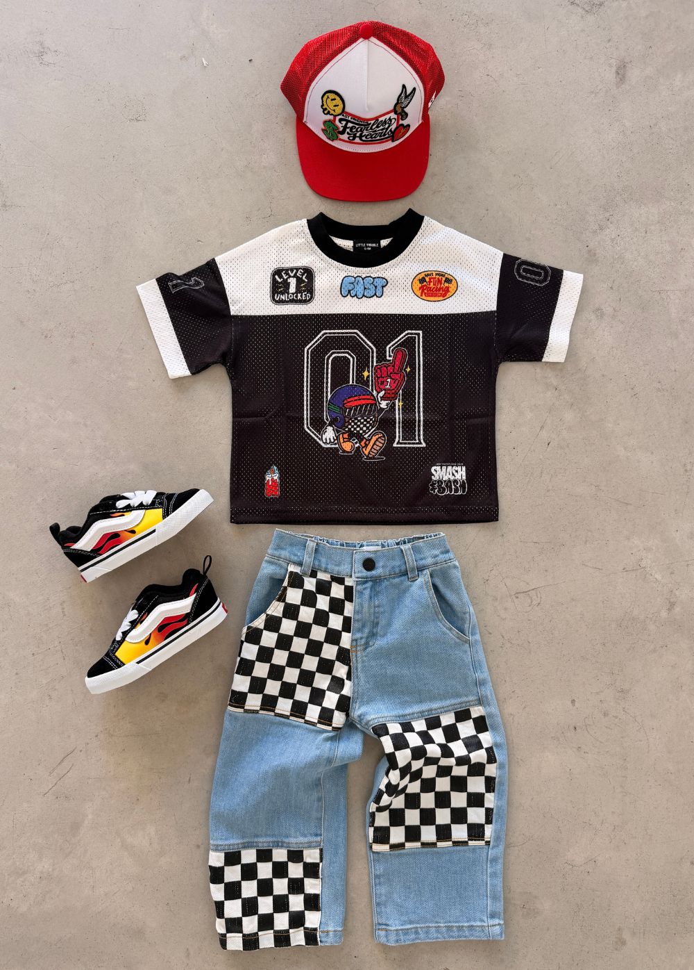 A children's birthday outfit set laid flat on a light concrete surface, including a black and white mesh jersey featuring a large number 1 graphic, jeans, a red and white baseball cap, and black sneakers with yellow and white accents.