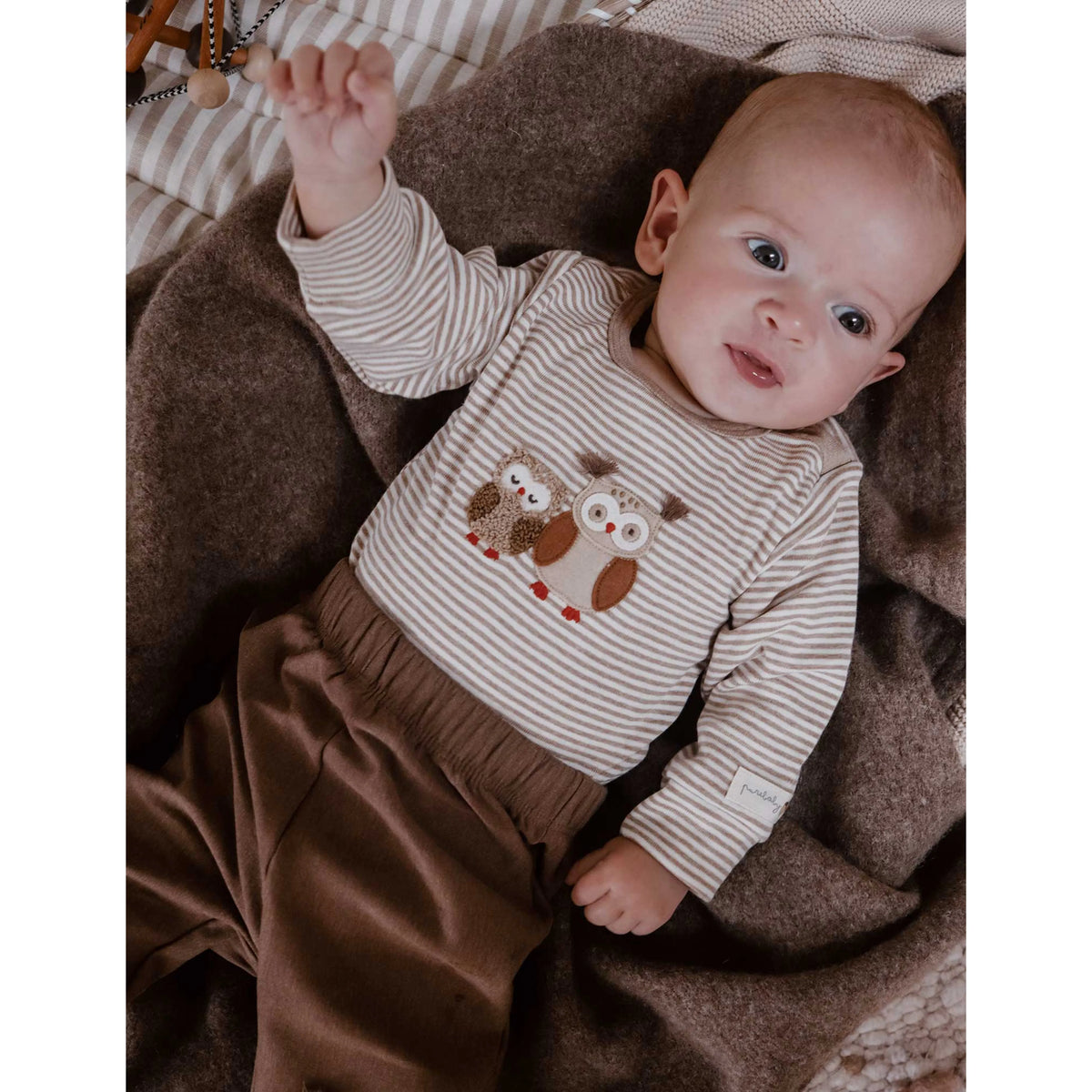 A baby lying on a brown blanket wearing a beige and white striped long-sleeve top with an owl graphic on the chest, paired with brown pants.