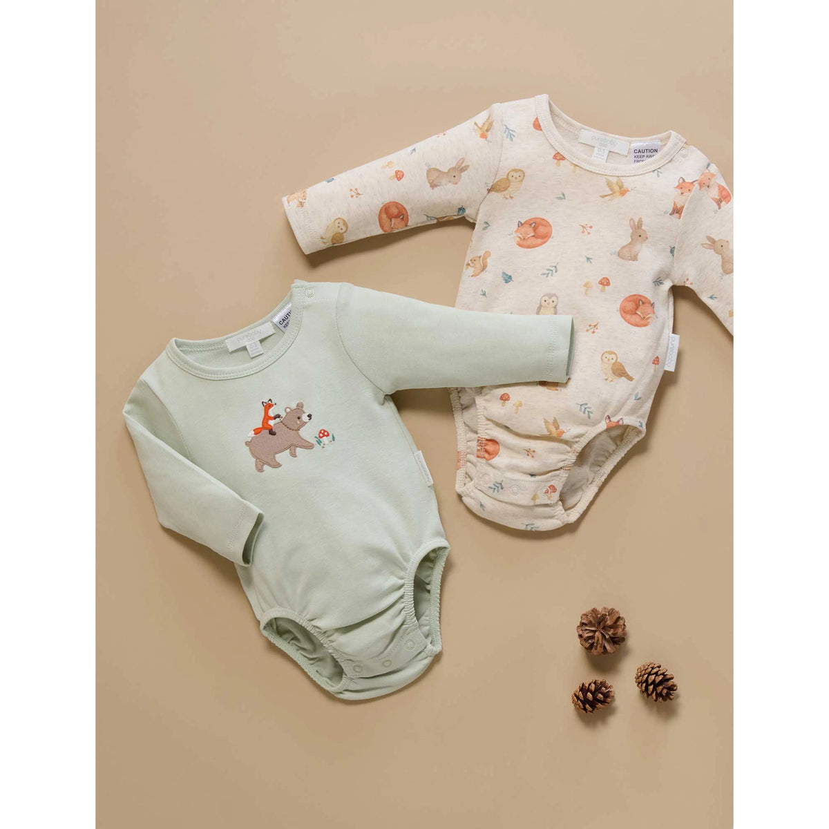 Two long-sleeve baby bodysuits laid flat on a beige background. One bodysuit is pale sage green with a small brown bear graphic on the chest.