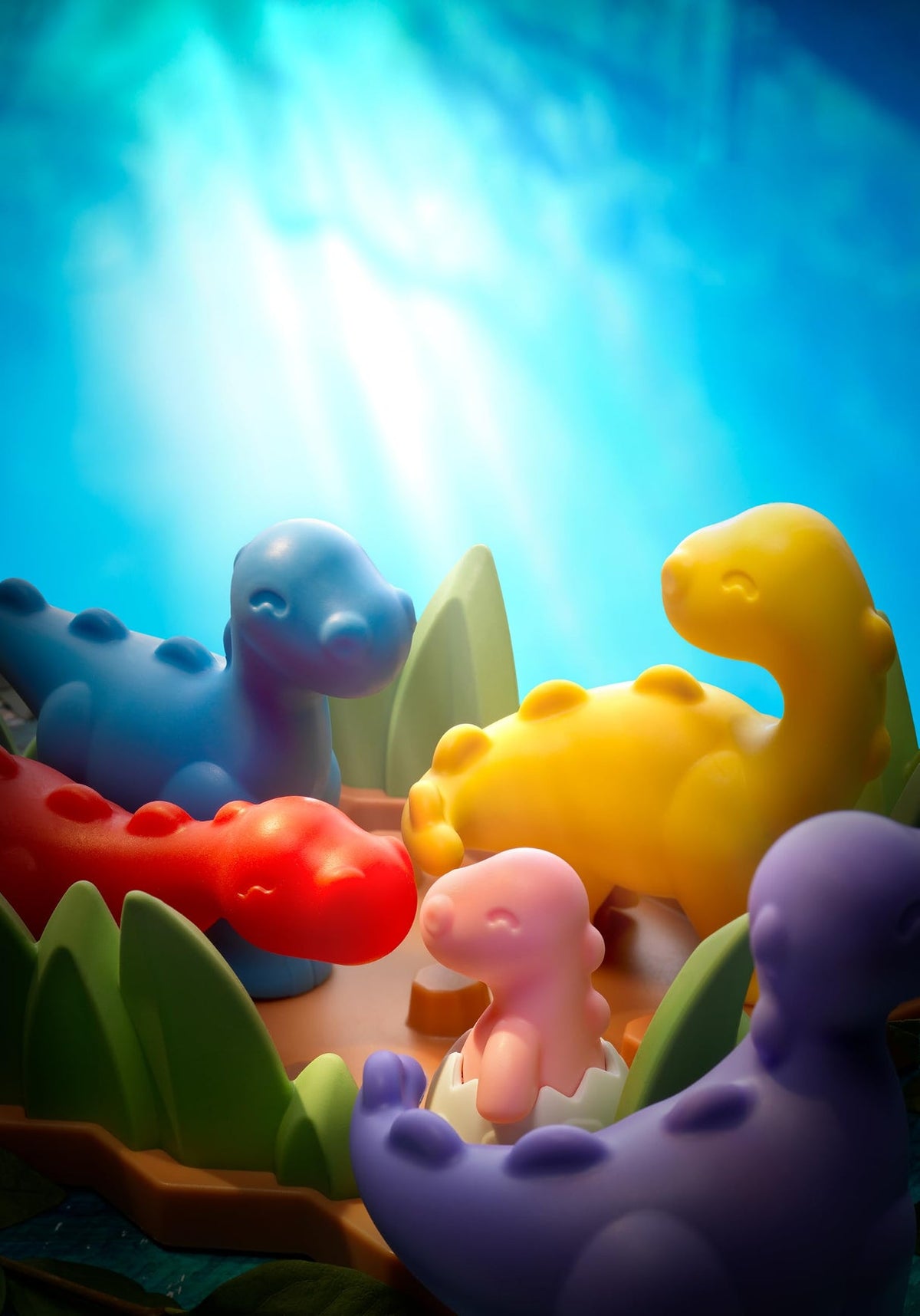 A colorful children's toy set featuring dinosaur figures and eggs on a brown base with green grass, photographed on a bright blue studio background.