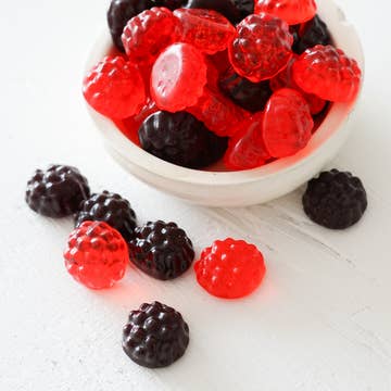 Gummy berries in red and black on a white surface with a bowl of gummy berries.