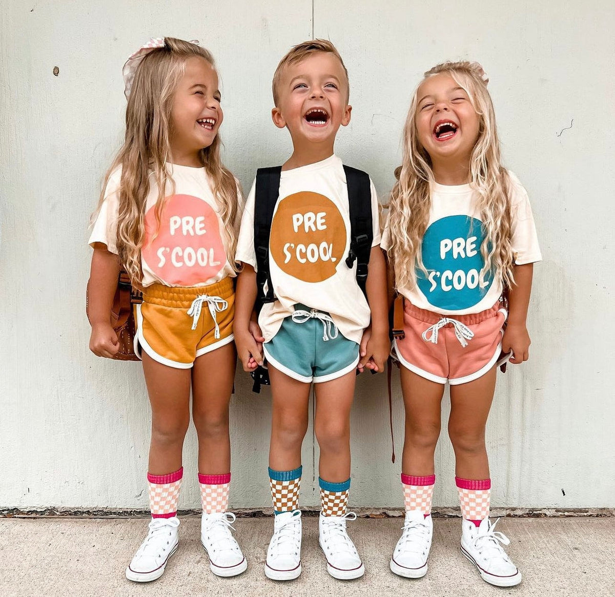 Three children standing together and laughing, wearing matching graphic t-shirts with colorful circular designs that read 'PRE S COOL' paired with different colored shorts, socks, and white sneakers against a plain light wall.