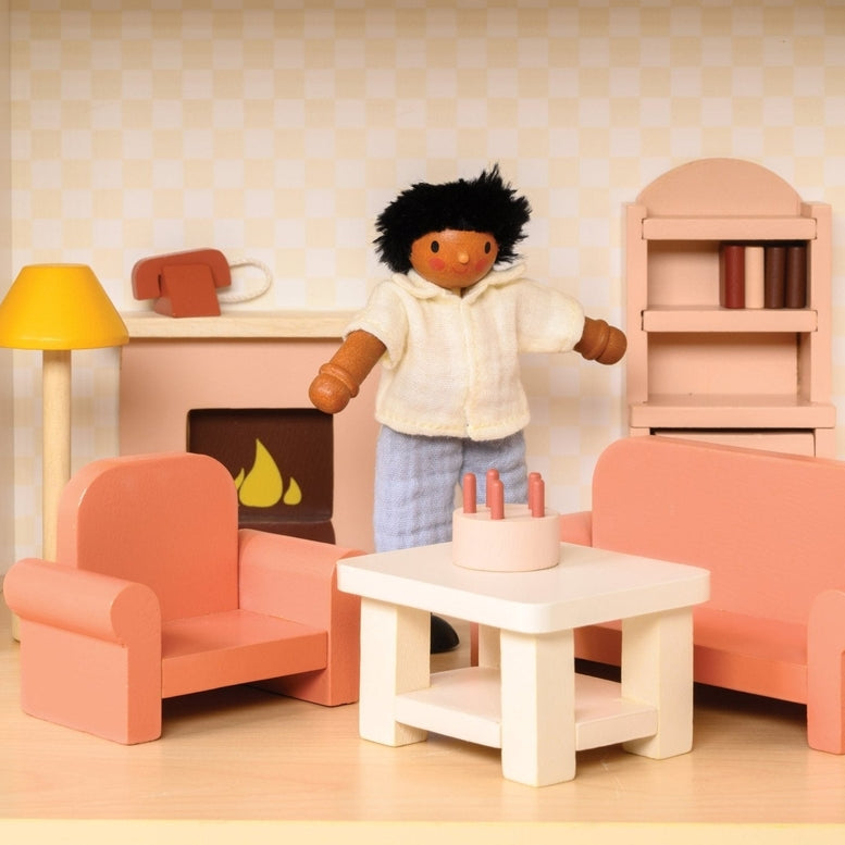 A miniature wooden dollhouse room interior featuring a wooden toy figure standing next to a coral-pink sofa and armchair set, a coffee table with a tiny red telephone, a pale pink decorative fireplace, a yellow floor lamp, and a natural wood bookcase against a patterned wallpaper background.
