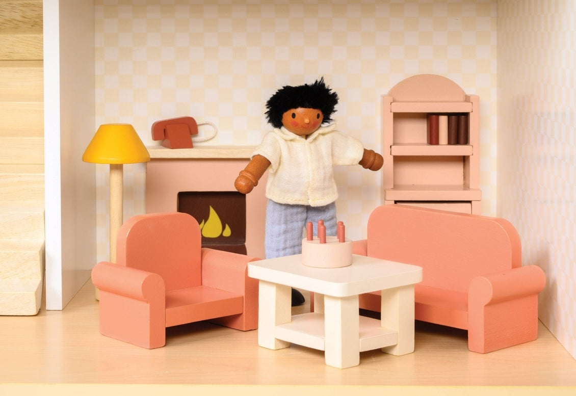 A miniature wooden dollhouse room interior featuring a wooden toy figure standing next to a coral-pink sofa and armchair set, a coffee table with a tiny red telephone, a pale pink decorative fireplace, a yellow floor lamp, and a natural wood bookcase against a patterned wallpaper background.