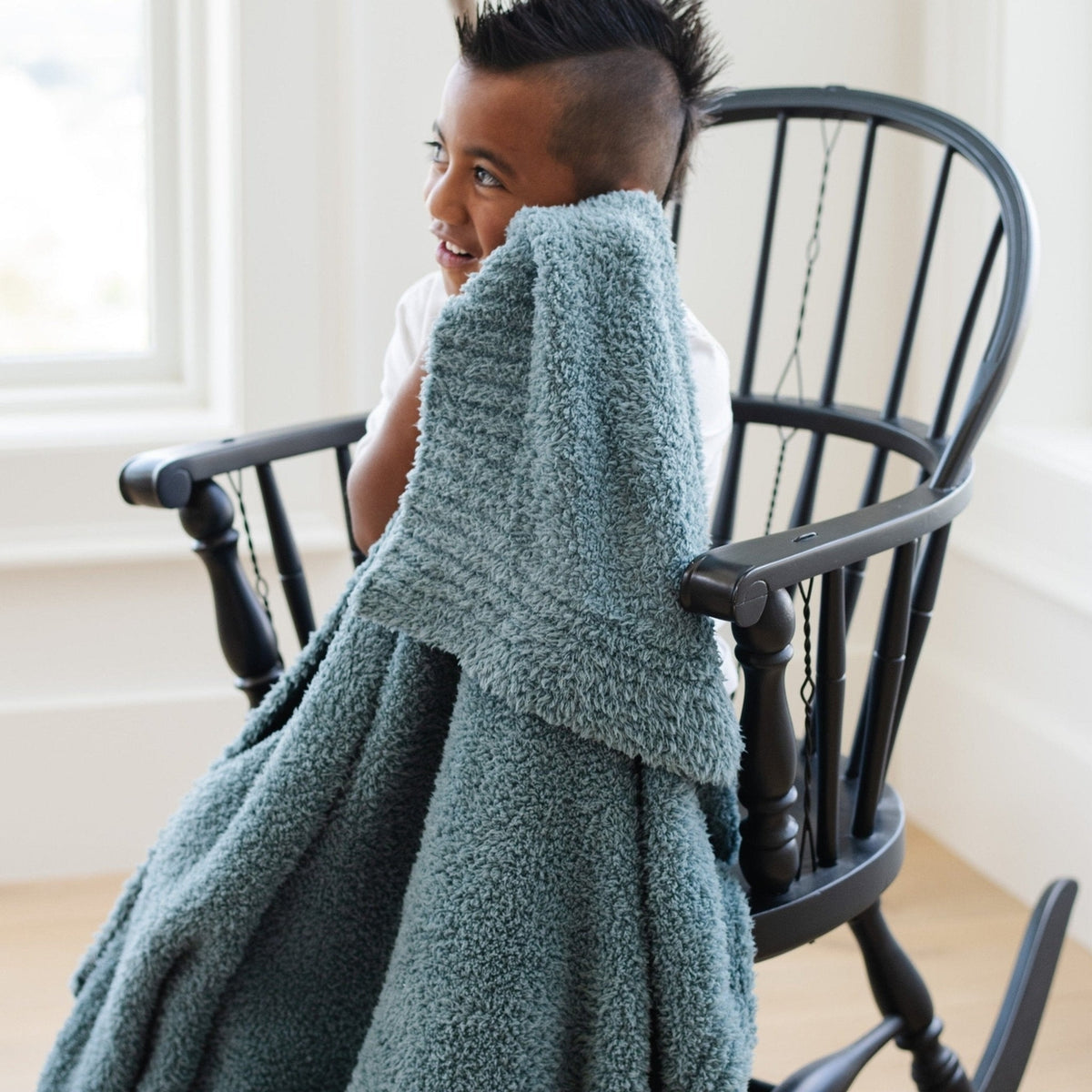Person sitting on a black chair with a green blanket draped over them, in a bright room.