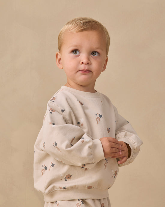 A toddler with blonde hair wearing a cream-colored long-sleeve sweatshirt with a small space-themed print of stars, planets, and rockets, posed against a neutral beige background.