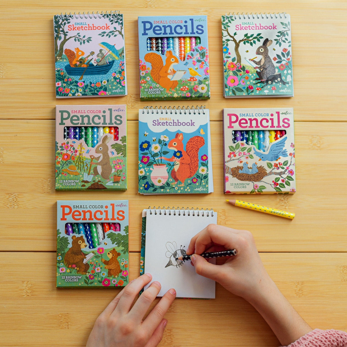 Set of art supplies including sketchbooks and colored pencils on a wooden surface with a hand holding a pencil.