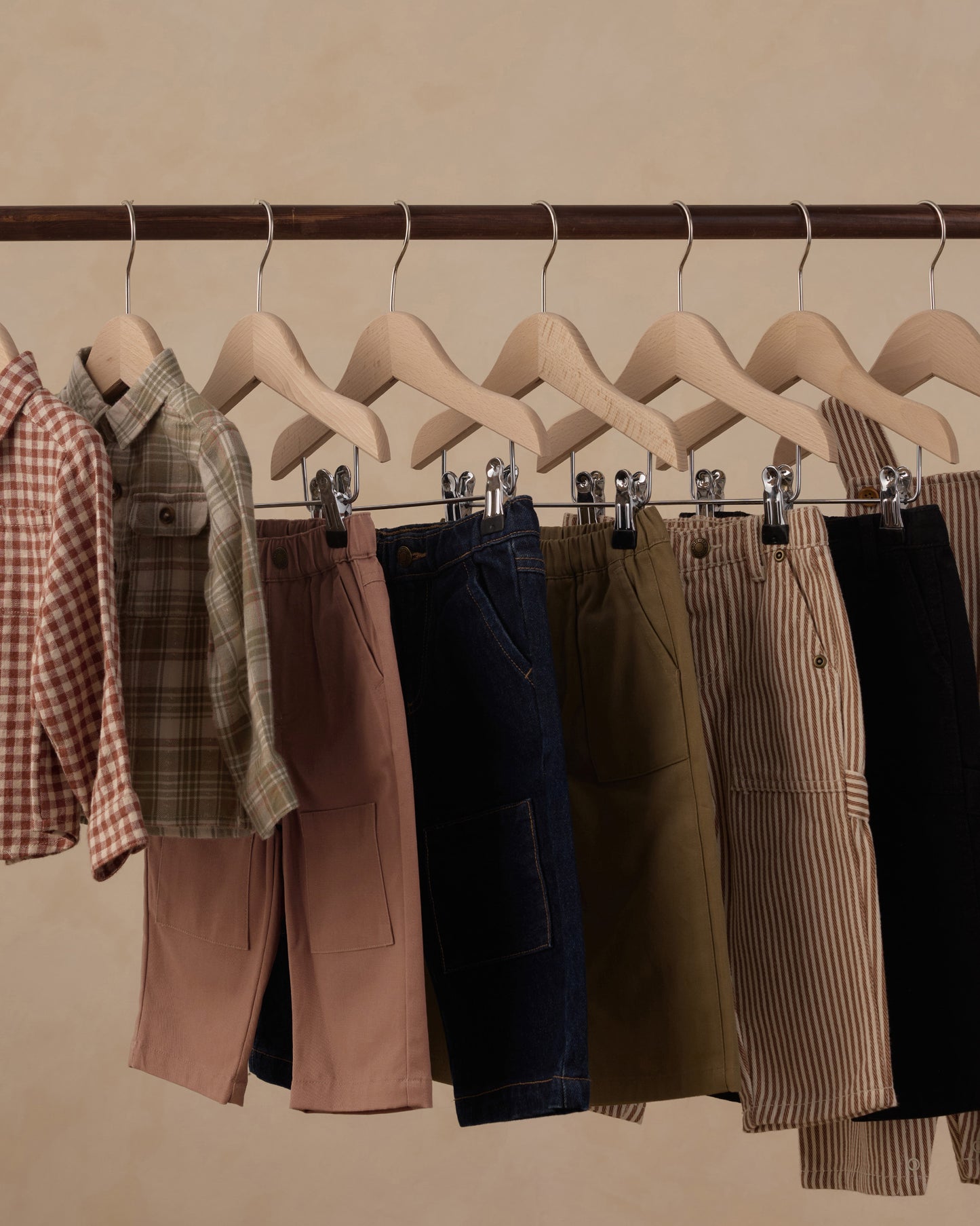A clothing rack displaying multiple pairs of shorts in different colors and patterns, including pink, navy, olive green, beige, and black, hanging on wooden hangers against a warm beige background.