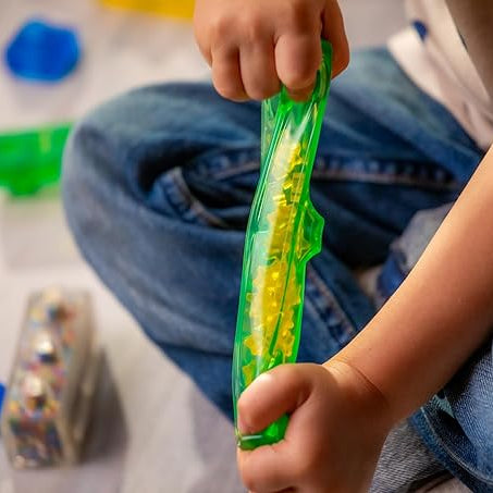 A child squeezes a long, translucent green Jelly Blox sensory building piece filled with small yellow beads, while other colorful blocks sit nearby on the floor.