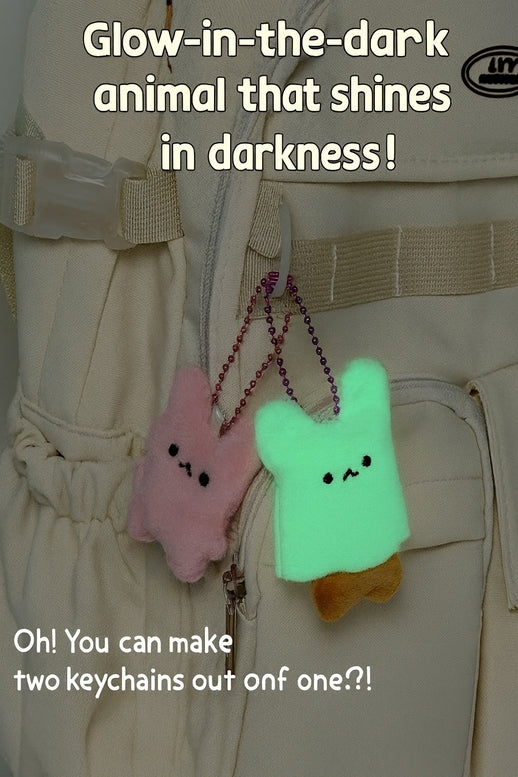 Two plush glow-in-the-dark keychains hanging from a beige utility belt. One keychain is pink and the other is glowing bright green, with a small brown accent on the green one.