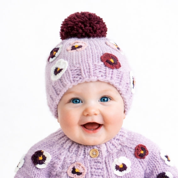 Baby wearing a purple knit outfit with floral patterns and a maroon pom-pom hat on a white background