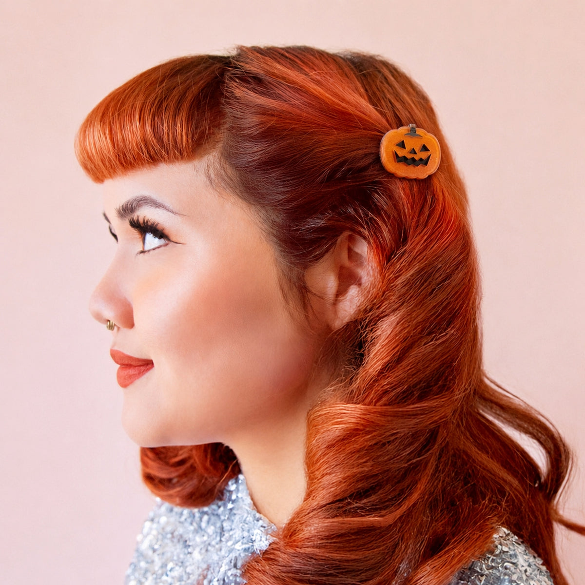 A woman in profile with bright red hair wearing a small orange pumpkin-shaped hair clip against a soft pink background.