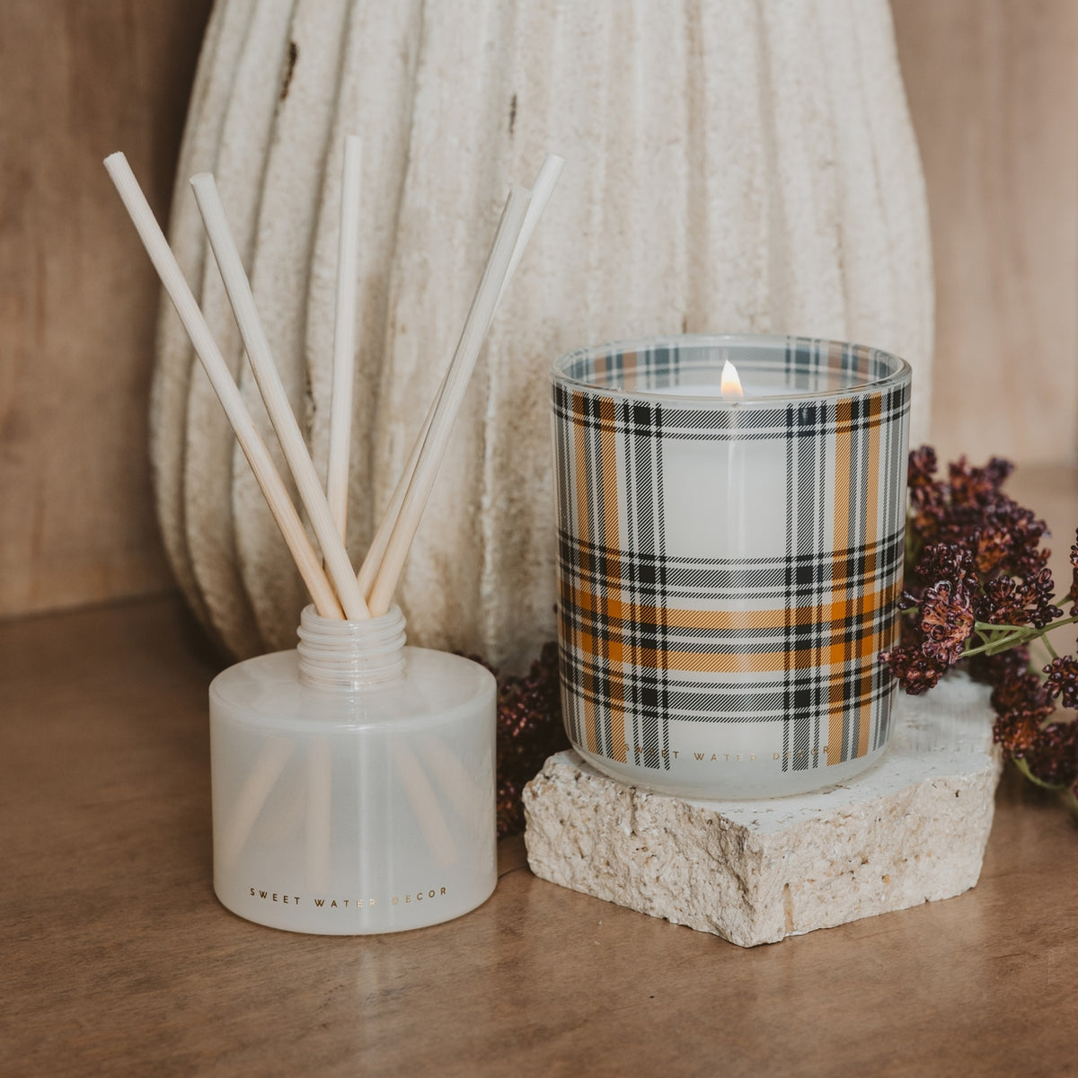 A reed diffuser bottle with white reeds and a lit glass candle in a plaid pattern, styled on a wooden surface with dried flowers and a textured cream vase in the background.