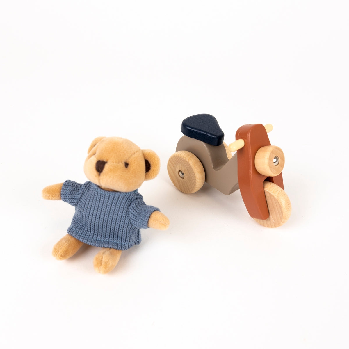 Small teddy bear in a blue dress next to a wooden toy vehicle on a white background