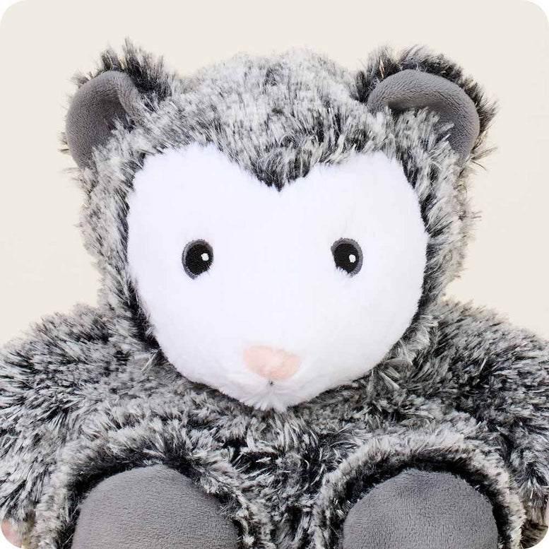 A close-up view of the Opossum by Warmies stuffed animal, highlighting its white face, round black button eyes, pink nose, and fluffy gray-and-black textured fur.