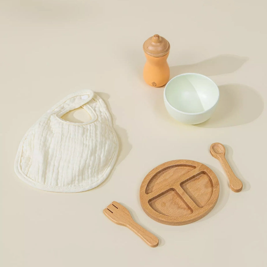 A baby feeding set arranged on a warm beige background, including a white muslin bib, a light wood divided plate, wooden utensils, a small white bowl, and an orange-toned bottle or dispenser.