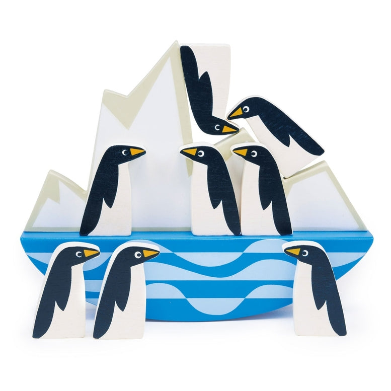 A collection of eight smooth, stylized wooden penguins stacked and balancing on a rocking white iceberg base, which rests on a blue-and-white wavy ocean design against a clean white background.