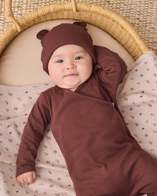A baby lying on a woven bassinet, wearing a ribbed brown long-sleeve sleep sack and matching hat with bear ears.