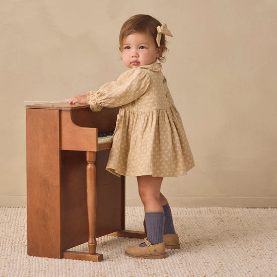 A toddler girl wearing a beige and white patterned long-sleeve dress, blue socks, and tan shoes stands next to a small wooden desk against a warm beige background.