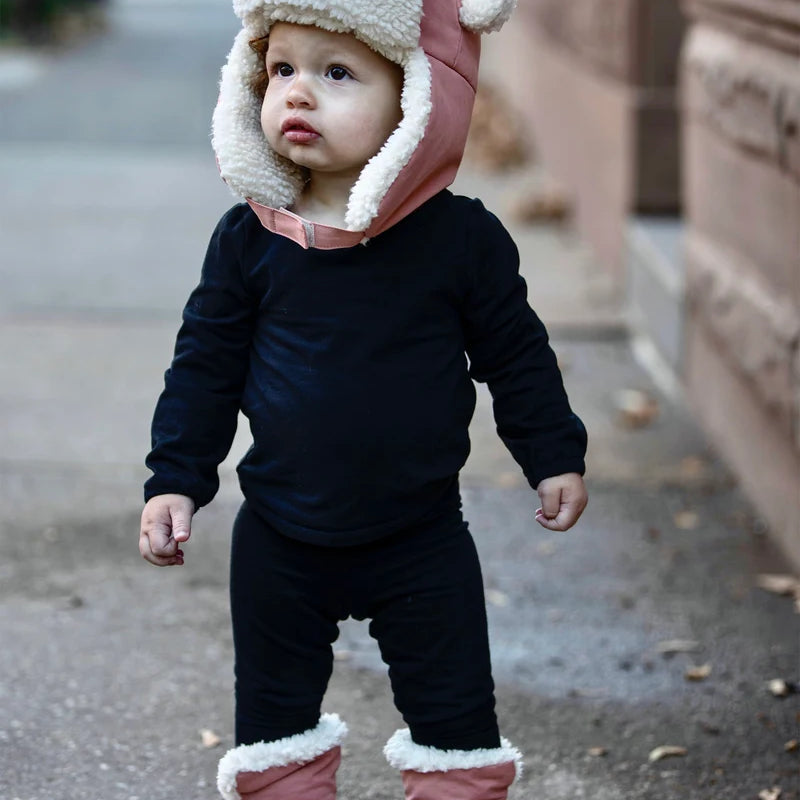 A toddler standing outdoors wearing a black outfit, pink winter boots with sherpa cuffs, and a pink and white sherpa-lined hat with bear ears.