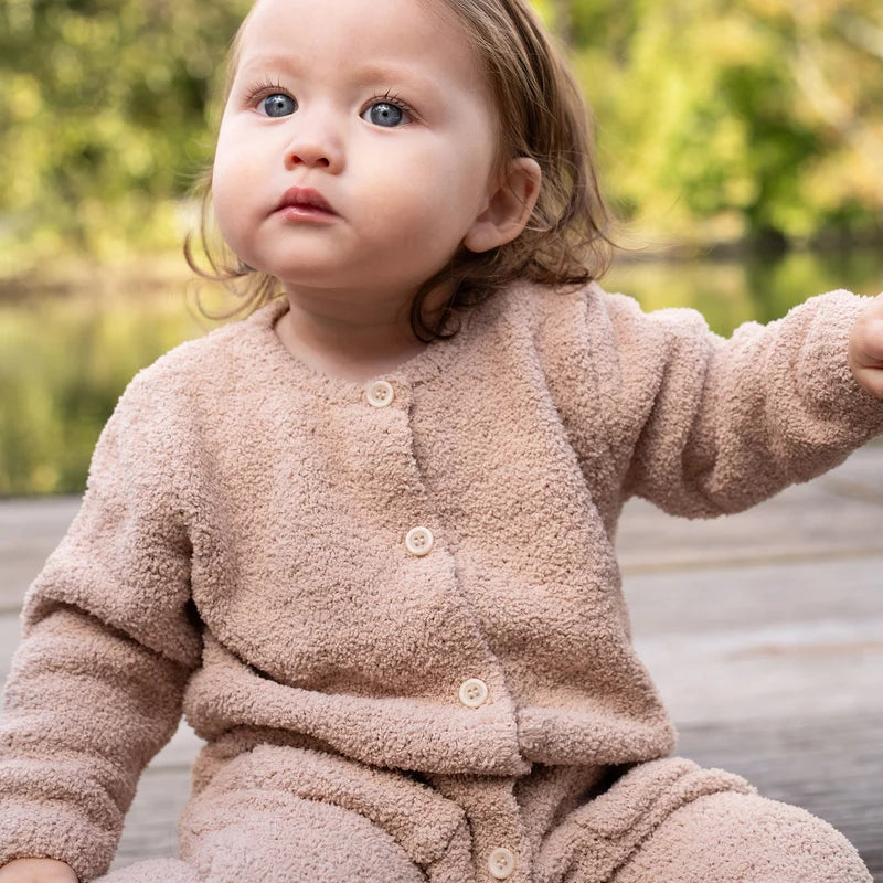 A toddler with reddish-brown hair sits outdoors in a natural, wooded setting, wearing a long-sleeve fuzzy romper in a warm pecan brown color by 7AM Enfant.