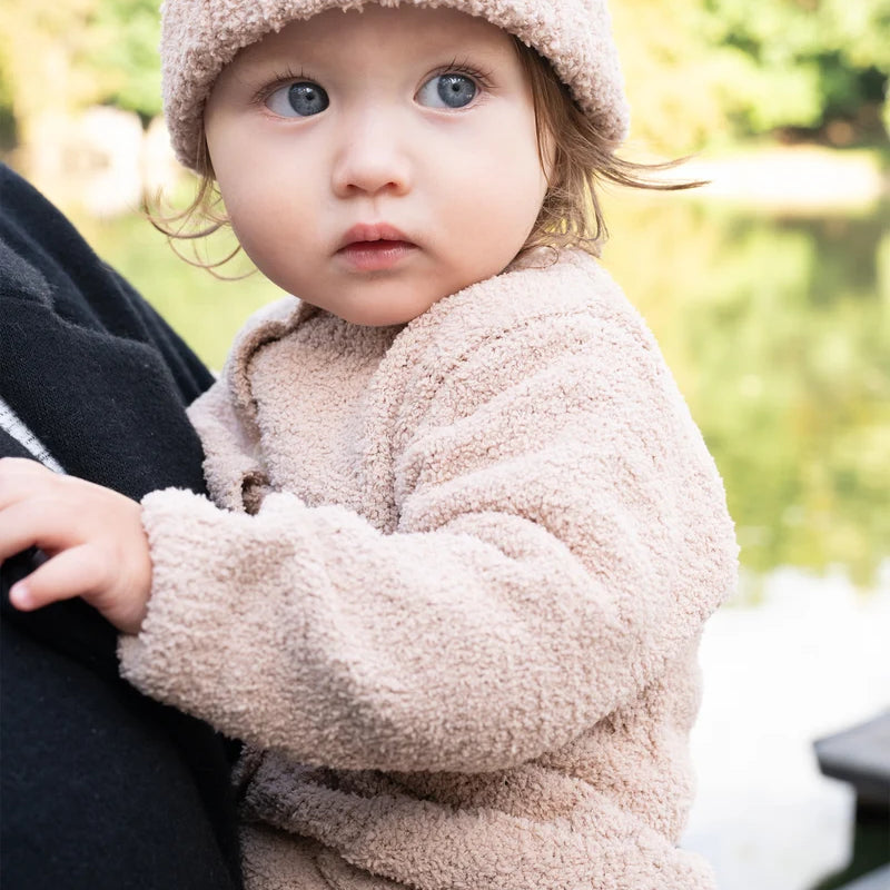 A toddler wearing a matching fuzzy cap and a long-sleeve pecan brown romper by 7AM Enfant is being held outdoors.
