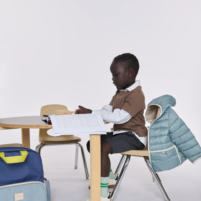 A young child sits at a small wooden desk in a bright studio setting, wearing a brown sweater over a white collared shirt, dark shorts, green socks, and white shoes. A light blue puffer jacket is draped over the chair behind them, and a blue-and-light-blue backpack rests on the floor to the left.