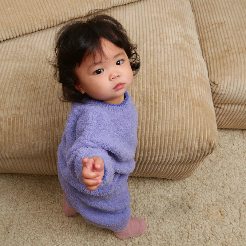 A toddler standing by a beige couch, wearing a soft, fuzzy knit taro purple sweater, pants, and a matching beanie.