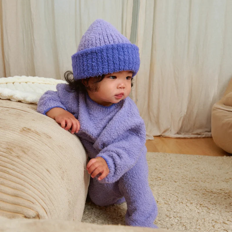 A baby standing by a beige couch, wearing a soft, fuzzy knit taro purple sweater, pants, and a matching beanie.