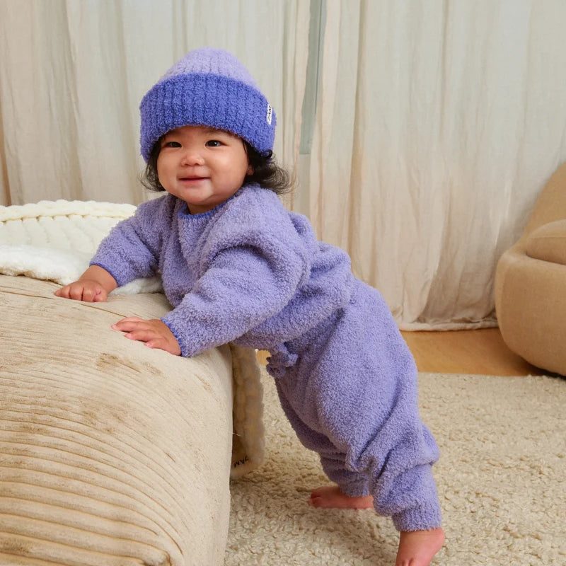 A baby standing by a beige couch, wearing a soft, fuzzy knit taro purple sweater, pants, and a matching beanie.