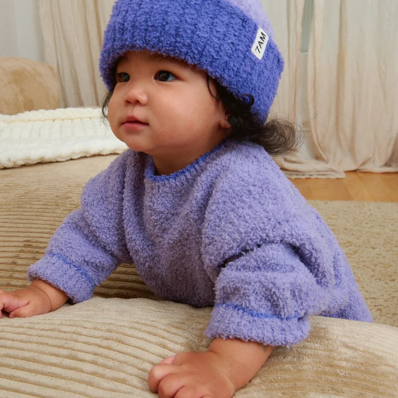 A toddler wearing a matching fuzzy purple knit sweater and a ribbed beanie with a folded brim and a white "7AM" tag.