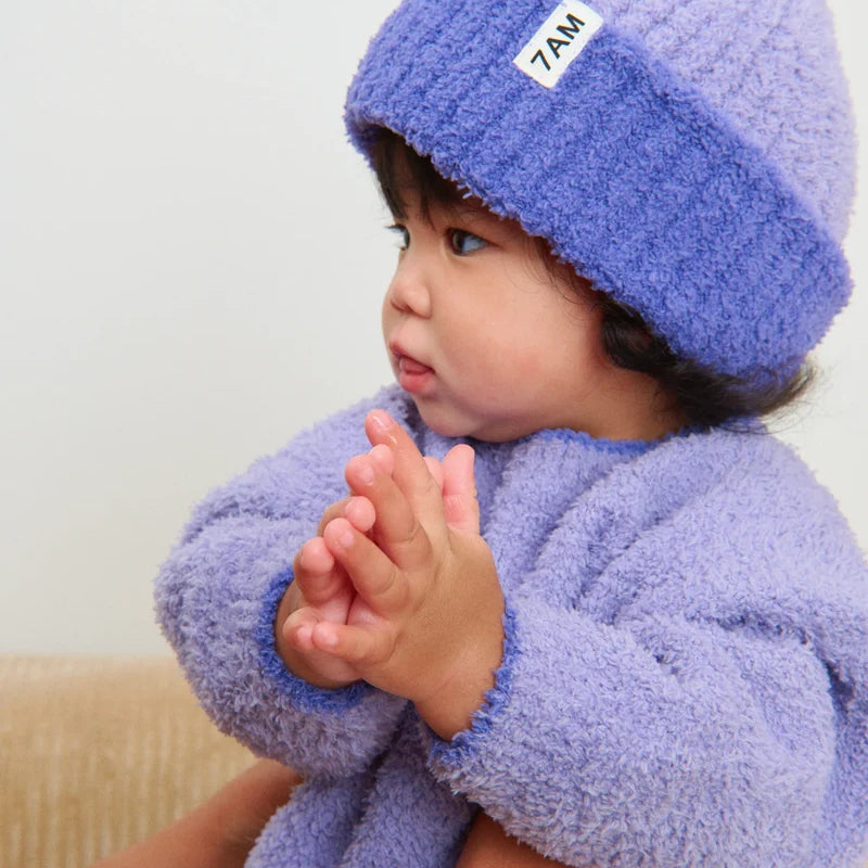 A toddler wearing a matching fuzzy purple knit sweater and a ribbed beanie with a folded brim and a white "7AM" tag.