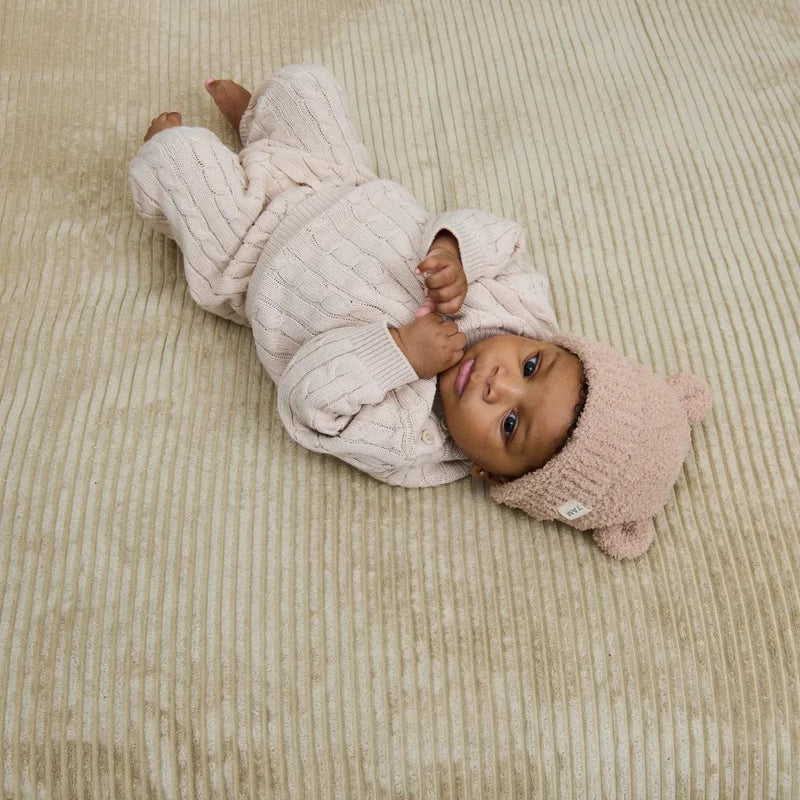 A baby lying on a textured neutral-colored blanket, wearing a matching ribbed cream-colored outfit and a fuzzy pecan-colored knit bear beanie with small ears.